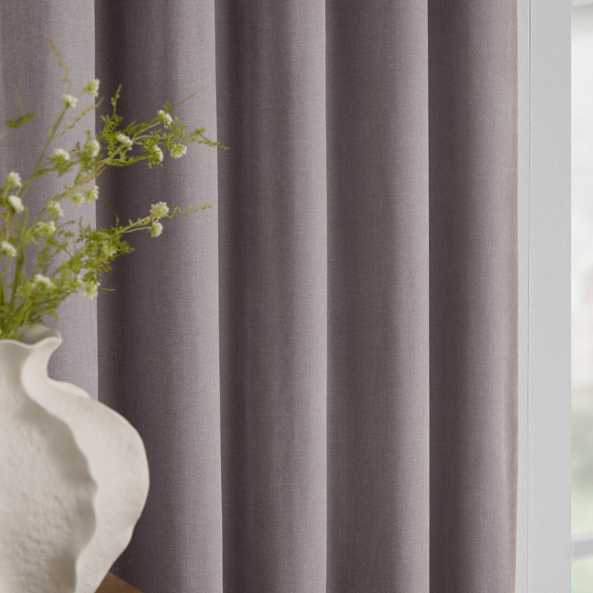 Close-up of mauve heavy chenille pencil pleat curtains with soft folds, shown beside a window with a vase of greenery in a living room setting.