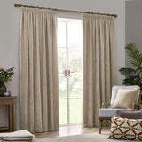 Natural beige pencil pleat curtains in heavy chenille velvet fabric, hung from a wood pole either side of a garden door in a living room with an armchair and cushions.