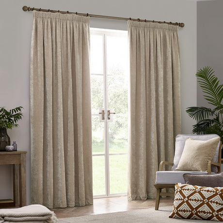 Natural beige pencil pleat curtains in heavy chenille velvet fabric, hung from a wood pole either side of a garden door in a living room with an armchair and cushions.