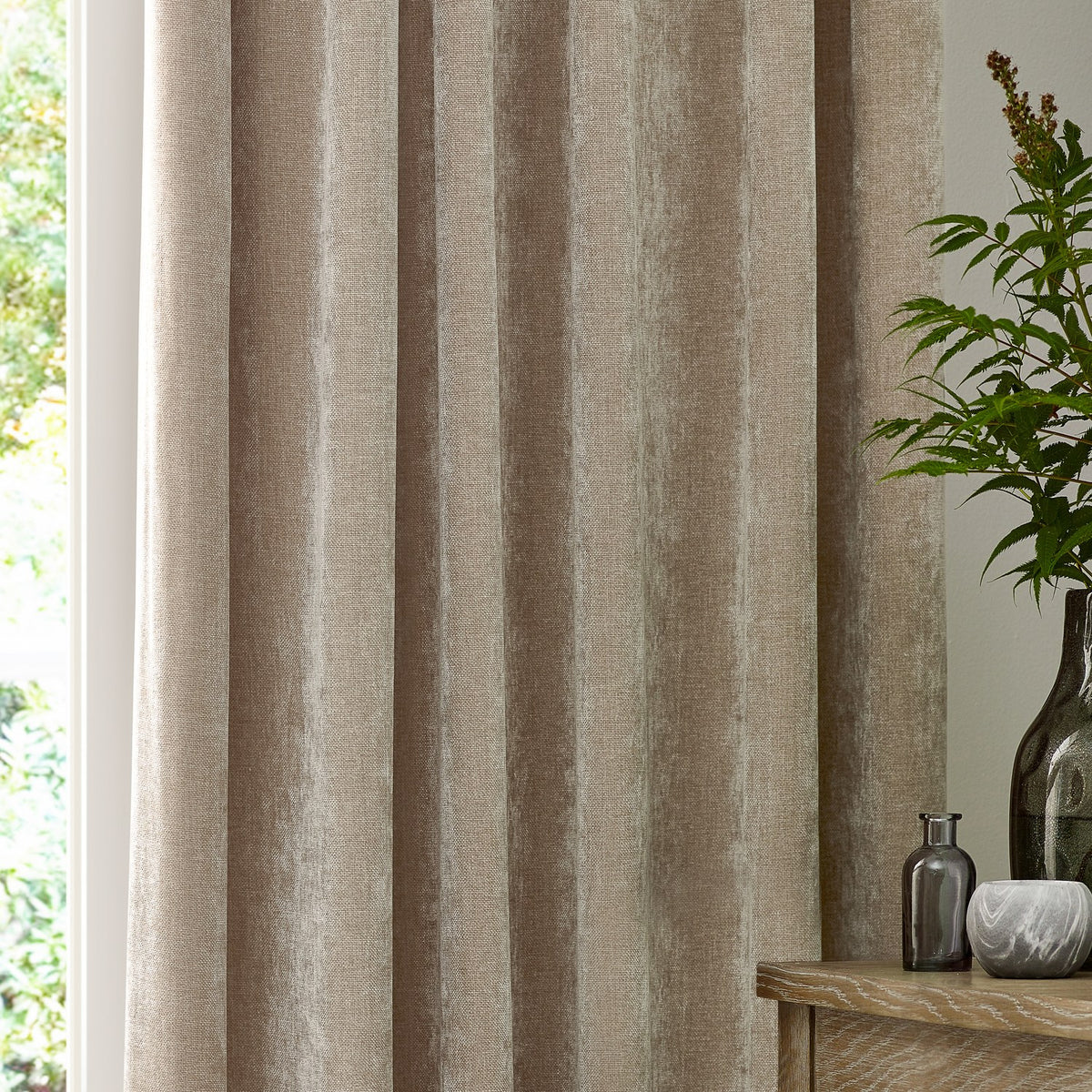 Heavy Chenille Plain Velvet Pencil Pleat Curtains in natural beige, shown hanging in soft folds beside a window in a living room setting with plants and decor nearby.