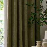 Heavy chenille velvet pencil pleat curtains in olive green, shown floor-length with soft folds beside a window in a living room setting.