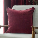 Square Heavy Chenille Plain Velvet Cushion in beetroot red with piped edge trim, styled on a light armchair beside a wooden armrest.