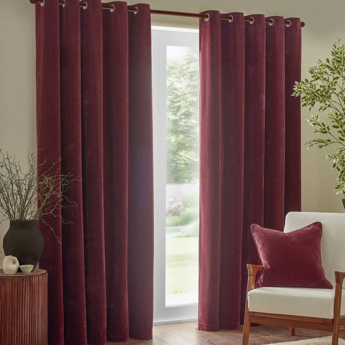 Square beetroot red velvet cushion with piped edge on a cream armchair, styled with burgundy curtains in a living room setting.