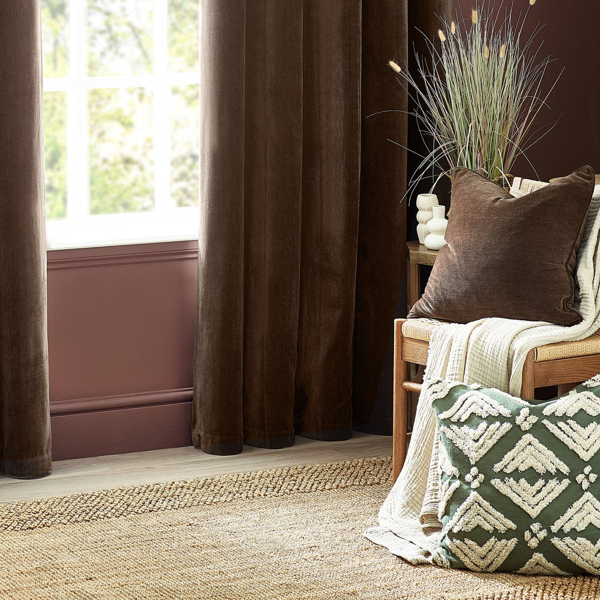 Square brown chenille velvet cushion on a wooden bench in a living room, styled with a green patterned cushion, cream throw, and dark curtains.