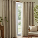 Square beige chenille cushion with piped trim edging, shown on a wooden armchair beside floor-length curtains in a living room setting.