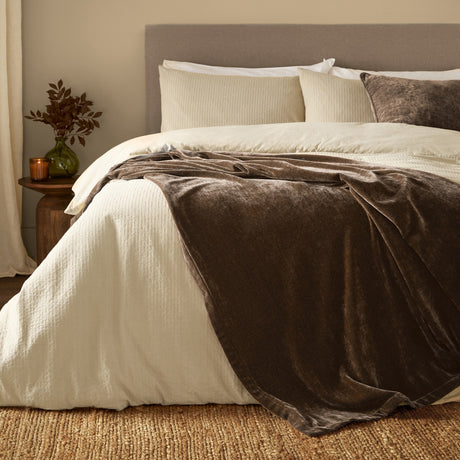 Brown velvet chenille throw draped over a bed with cream bedding, layered on cream pillows and a headboard in a bedroom setting.