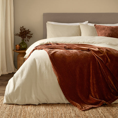 Nutmeg brown chenille velvet throw draped over a bed with cream bedding, layered pillows, and a rust velvet cushion in a bedroom setting.