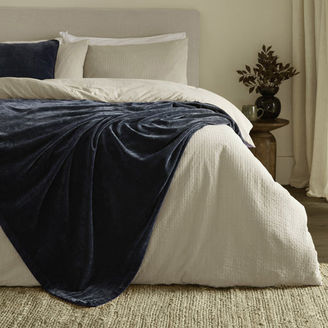 Navy velvet chenille throw draped over a bed with cream bedding, layered with pillows in a neutral bedroom setting.