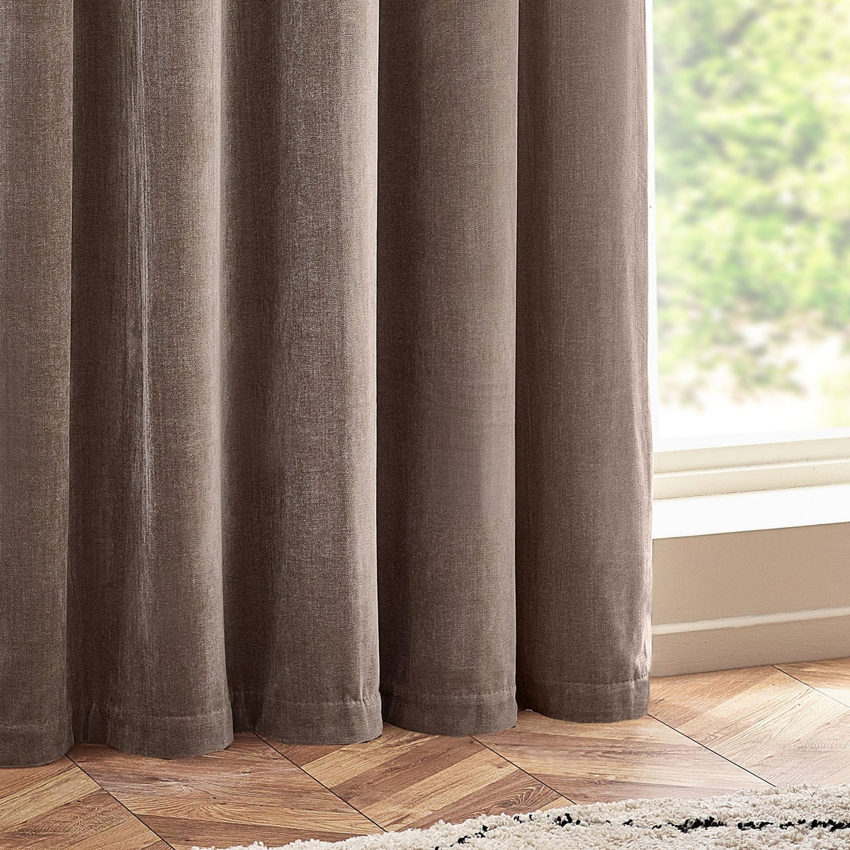 Heavy chenille velvet eyelet curtains in greige, shown draped in soft folds beside a bright window, with a herringbone wood floor and cream rug visible.