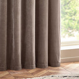 Heavy chenille velvet eyelet curtains in greige, shown draped in soft folds beside a bright window, with a herringbone wood floor and cream rug visible.