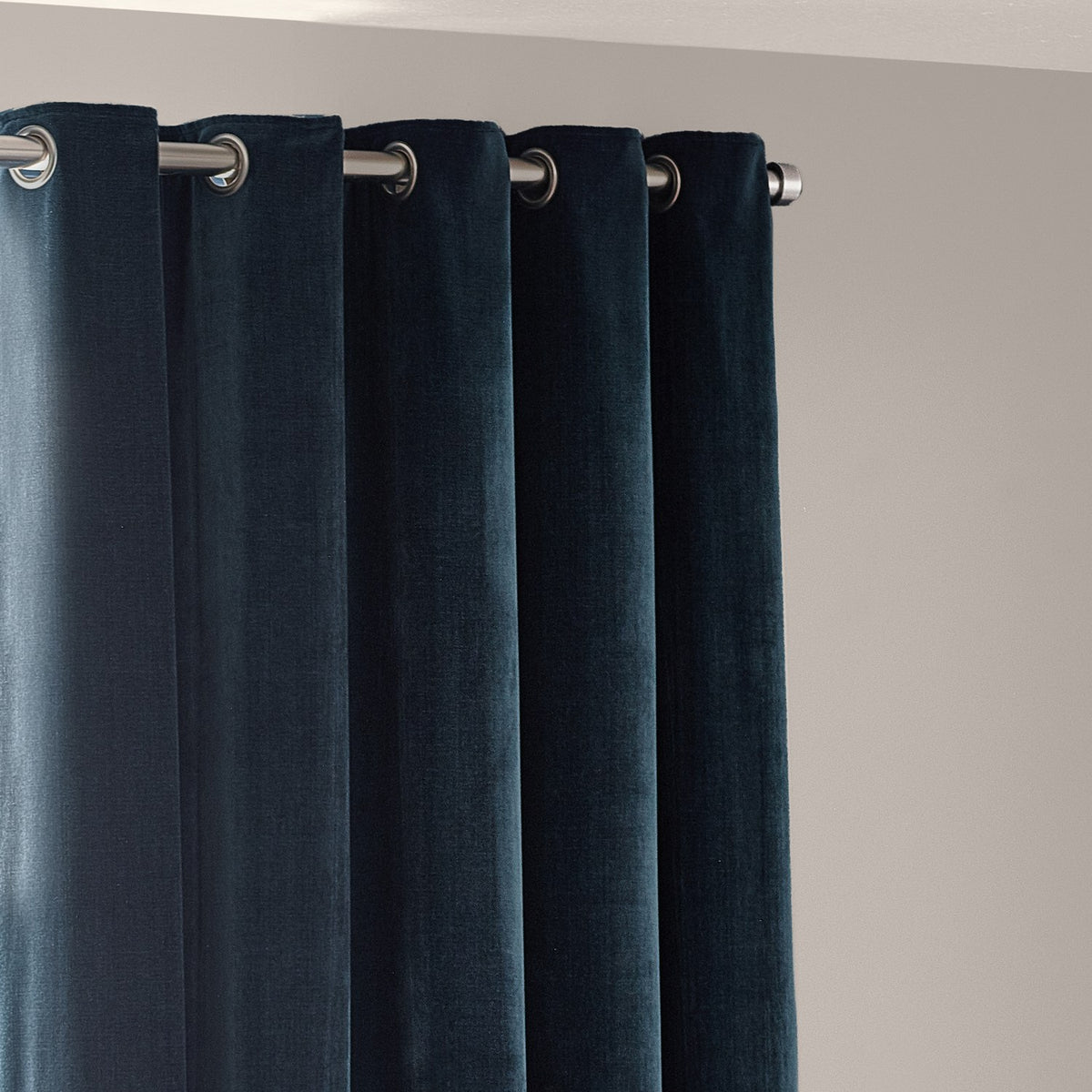 Heavy chenille velvet eyelet curtains in navy blue, hung on a chrome pole with metal grommets, falling in full rounded folds against a light wall.