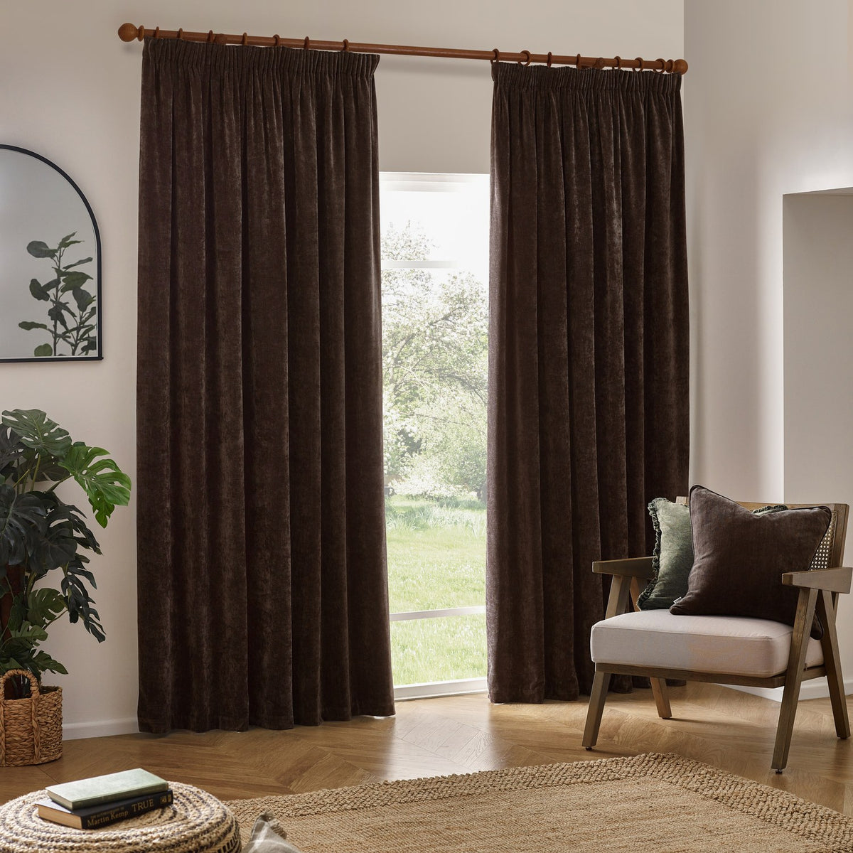 Brown heavy chenille velvet pencil pleat curtains hanging on a wooden pole beside a garden window in a living room with an armchair and cushions.