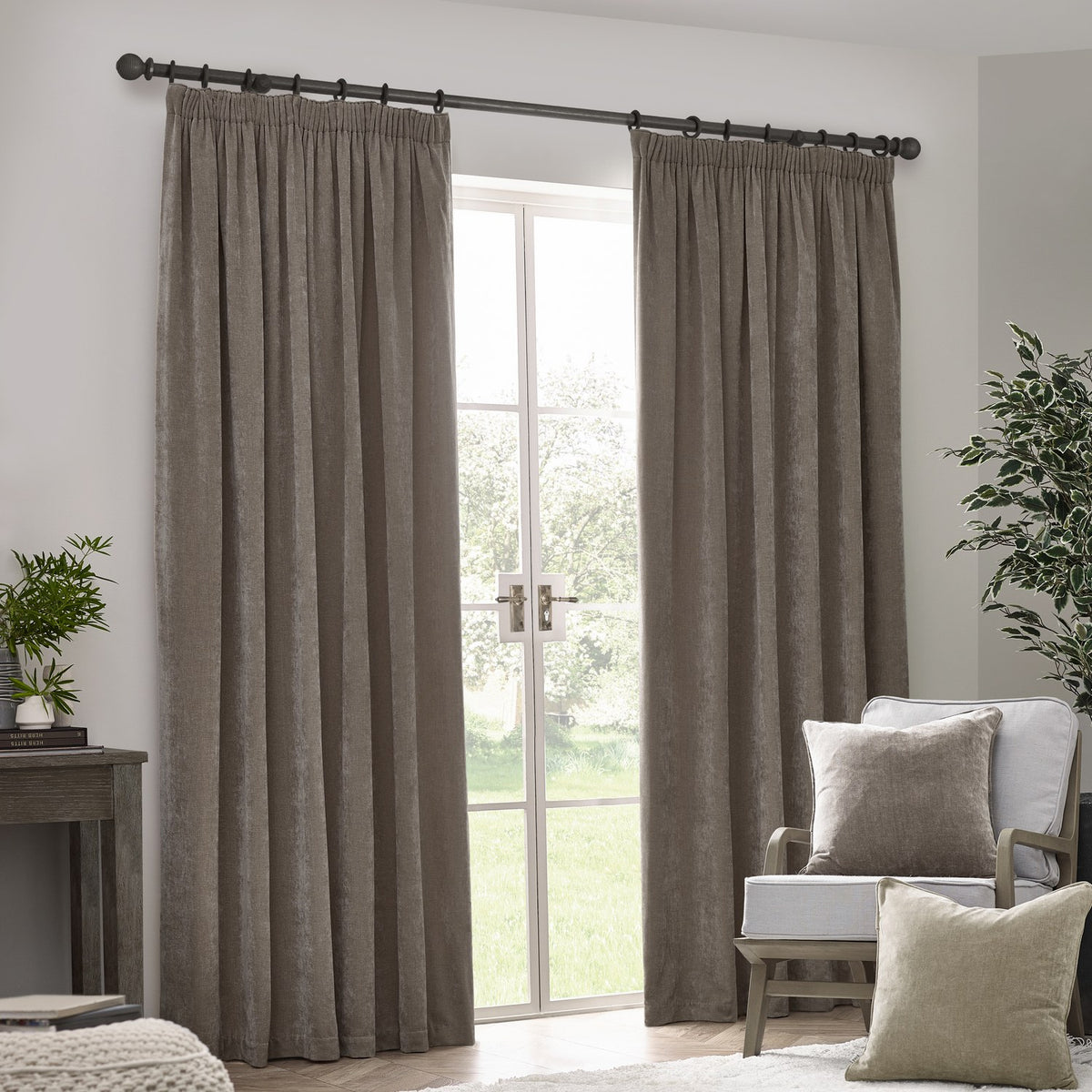 Heavy Chenille Plain Velvet Pencil Pleat Curtains in greige, shown hanging by a French door in a living room with an armchair and cushions nearby.