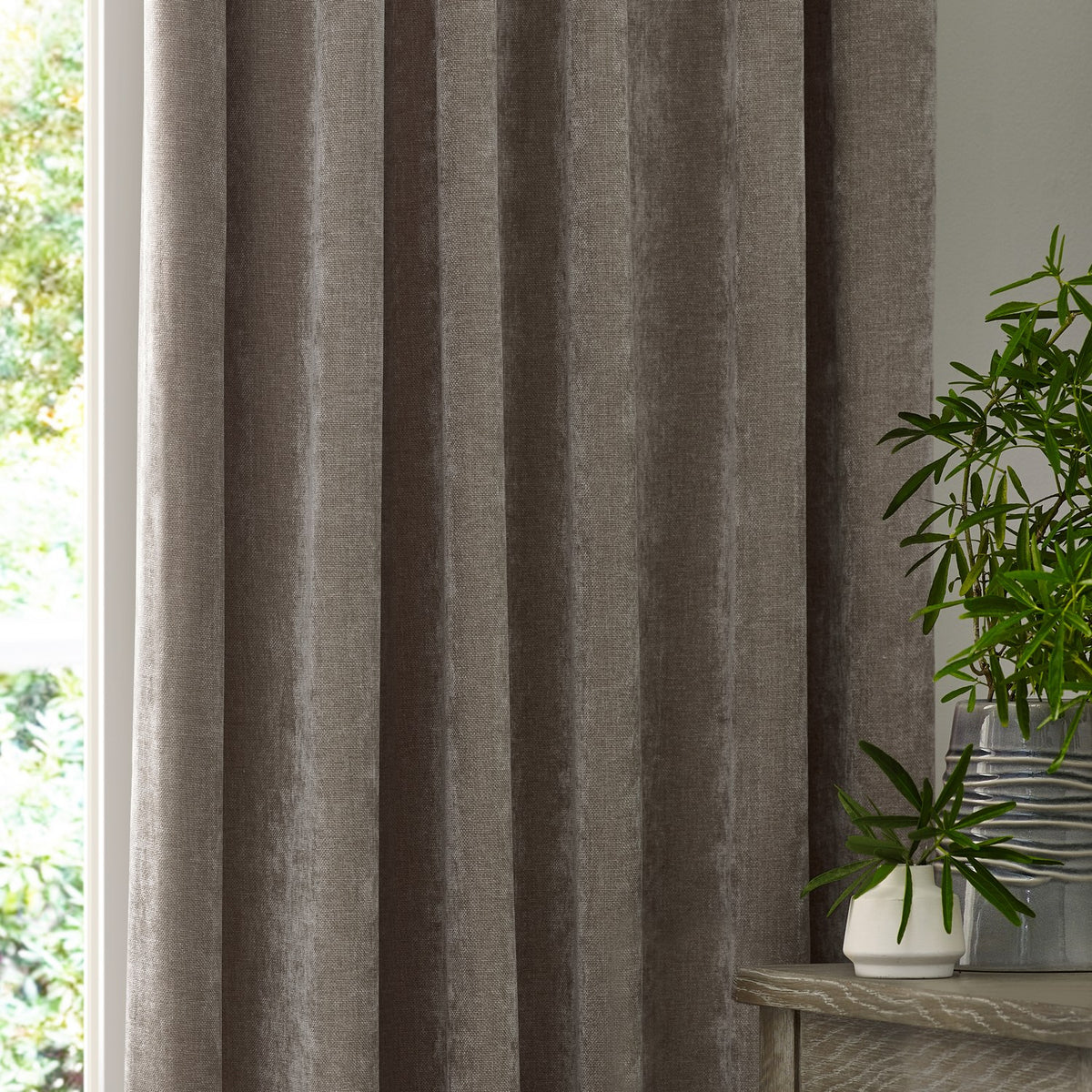 Heavy chenille velvet pencil pleat curtains in greige, shown hanging in soft folds beside a window, with plants and a wooden console table nearby.