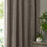 Heavy chenille velvet pencil pleat curtains in greige, shown hanging in soft folds beside a window, with plants and a wooden console table nearby.