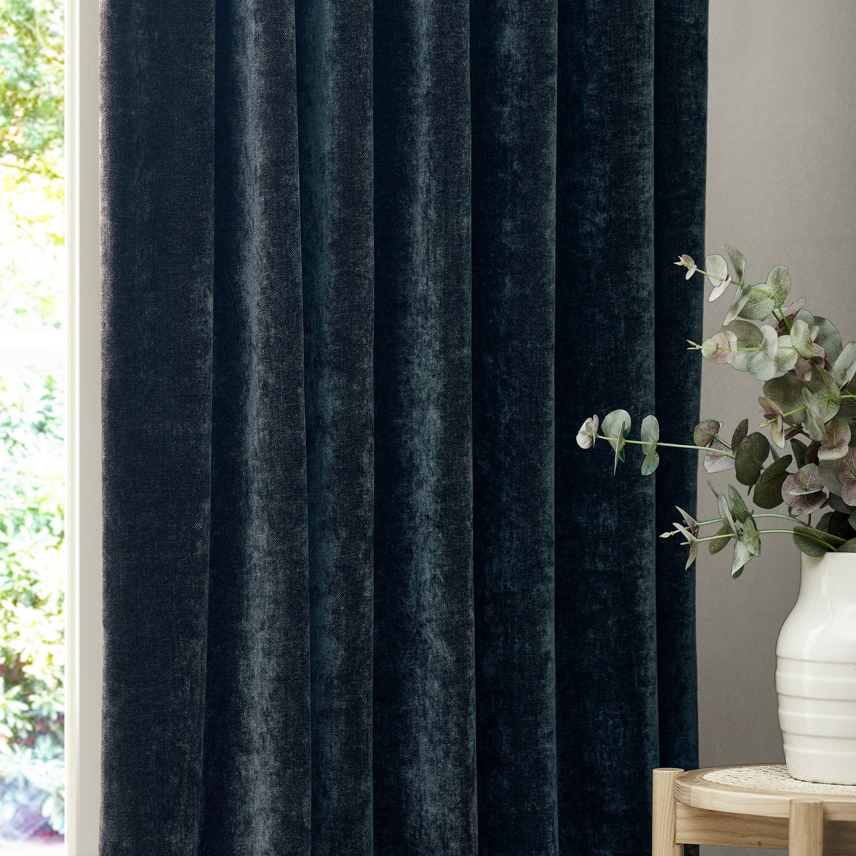Heavy chenille velvet pencil pleat curtains in navy blue, shown in deep folds by a window, with a eucalyptus plant in a white vase nearby.