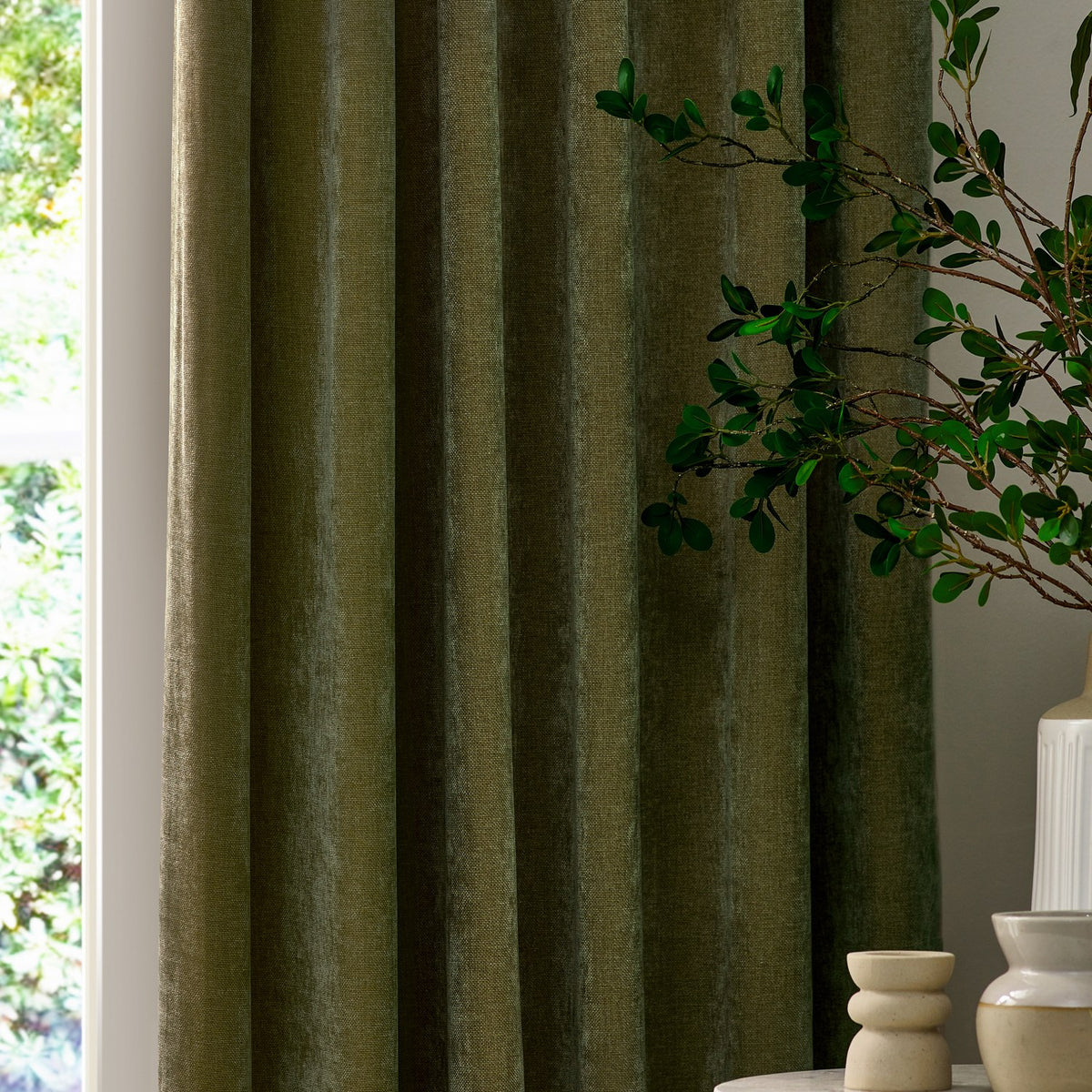 Heavy chenille velvet pencil pleat curtains in olive green, shown hanging in soft folds beside a window with greenery and decorative vases in a living room setting.