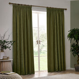 Olive green heavy chenille pencil pleat curtains hanging on a wooden pole beside a glazed door, shown in a living room with houseplants and wood flooring.