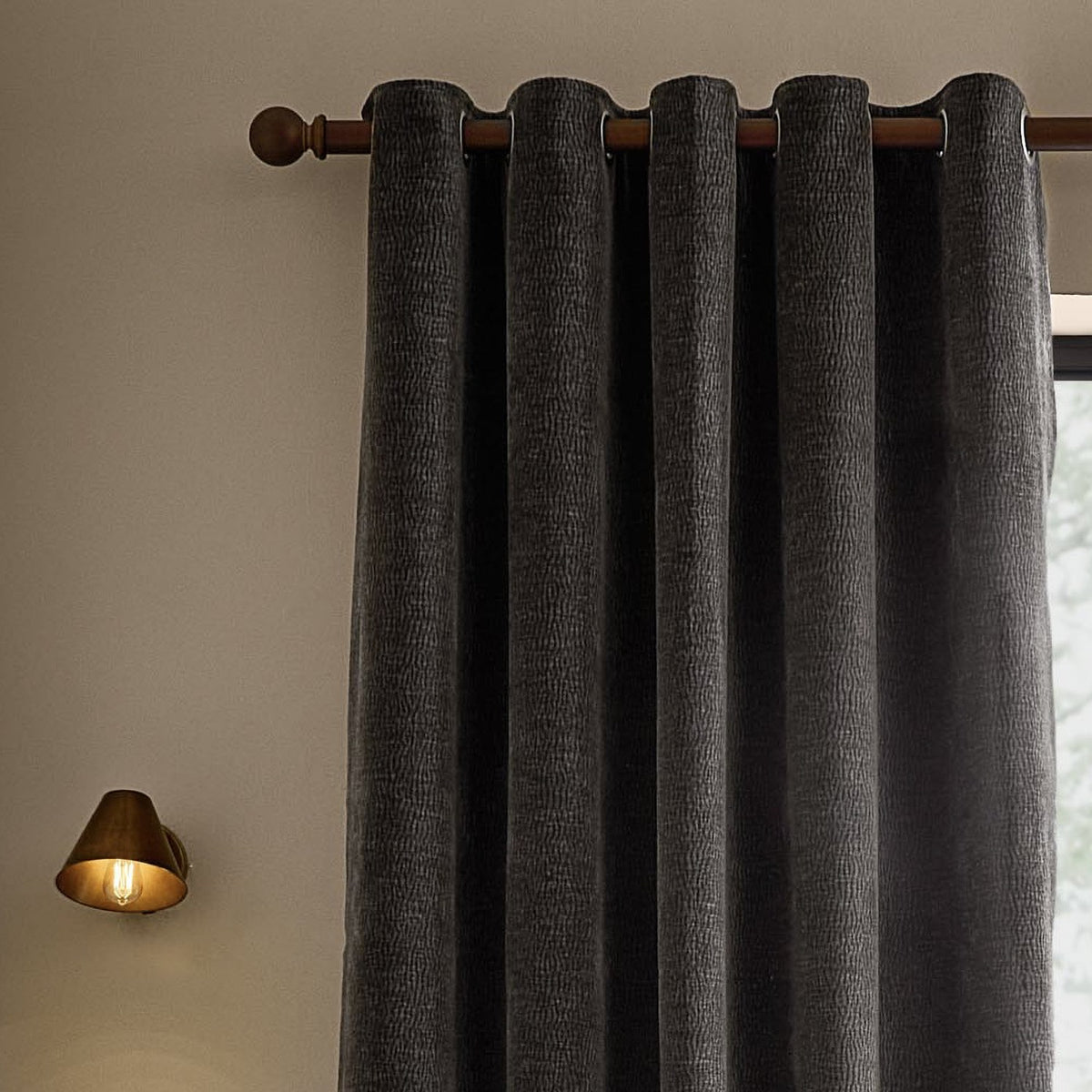 Heath Washed Velvet Fully Lined Eyelet Curtains in charcoal, textured jacquard fabric on a bronze eyelet rod, styled beside a brass wall light in a neutral-toned room.