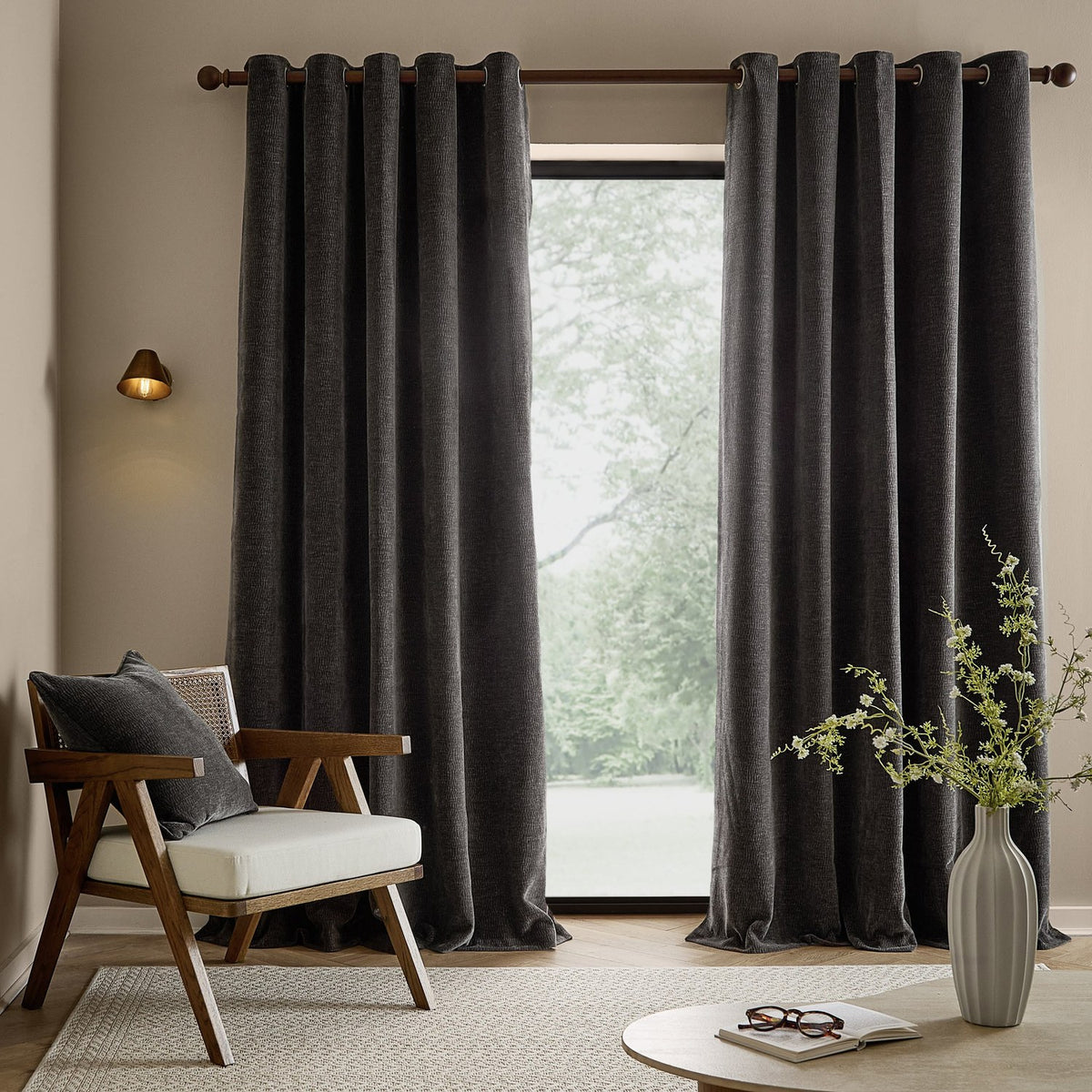 Heath Washed Velvet Fully Lined Eyelet Curtains in charcoal, shown in a living room with grommet top on a dark metal pole, framing a glass door beside a wooden armchair.