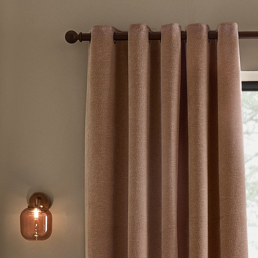 Heath Washed Velvet Jacquard Fully Lined Eyelet Curtains in Cotta pink, textured fabric on a dark rod, styled in a room beside a wall sconce and window.