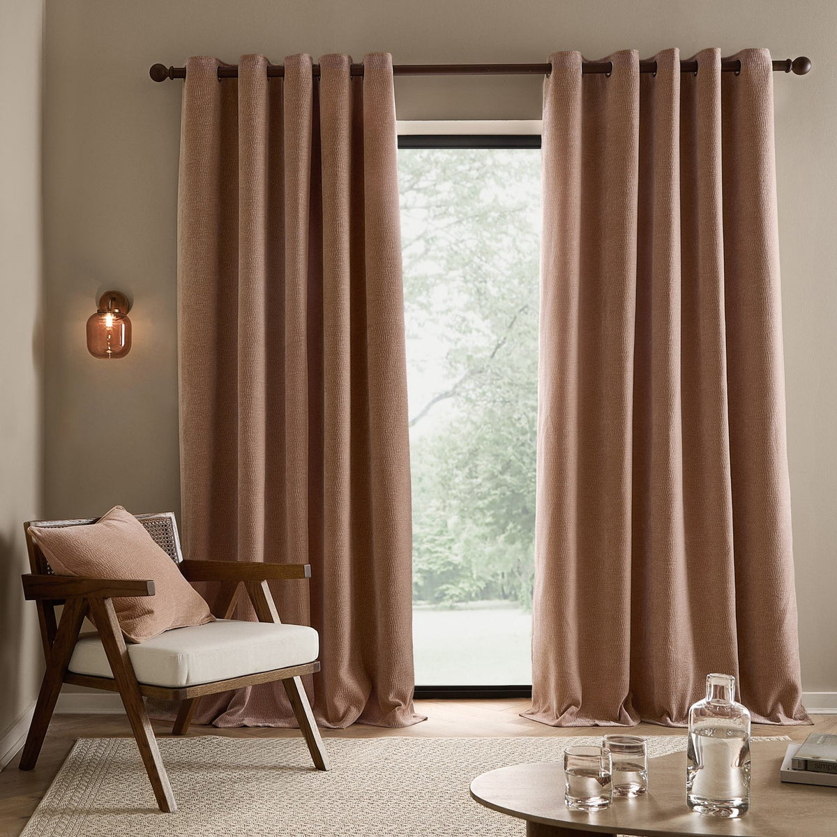 Heath Washed Velvet Jacquard fully lined eyelet curtains in dusky pink, hung on a dark rod beside a wooden armchair in a living room setting.