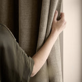 Heath Washed Velvet Jacquard Eyelet Curtains in greige, hand drawing back the textured, crinkled fabric panel beside a window.