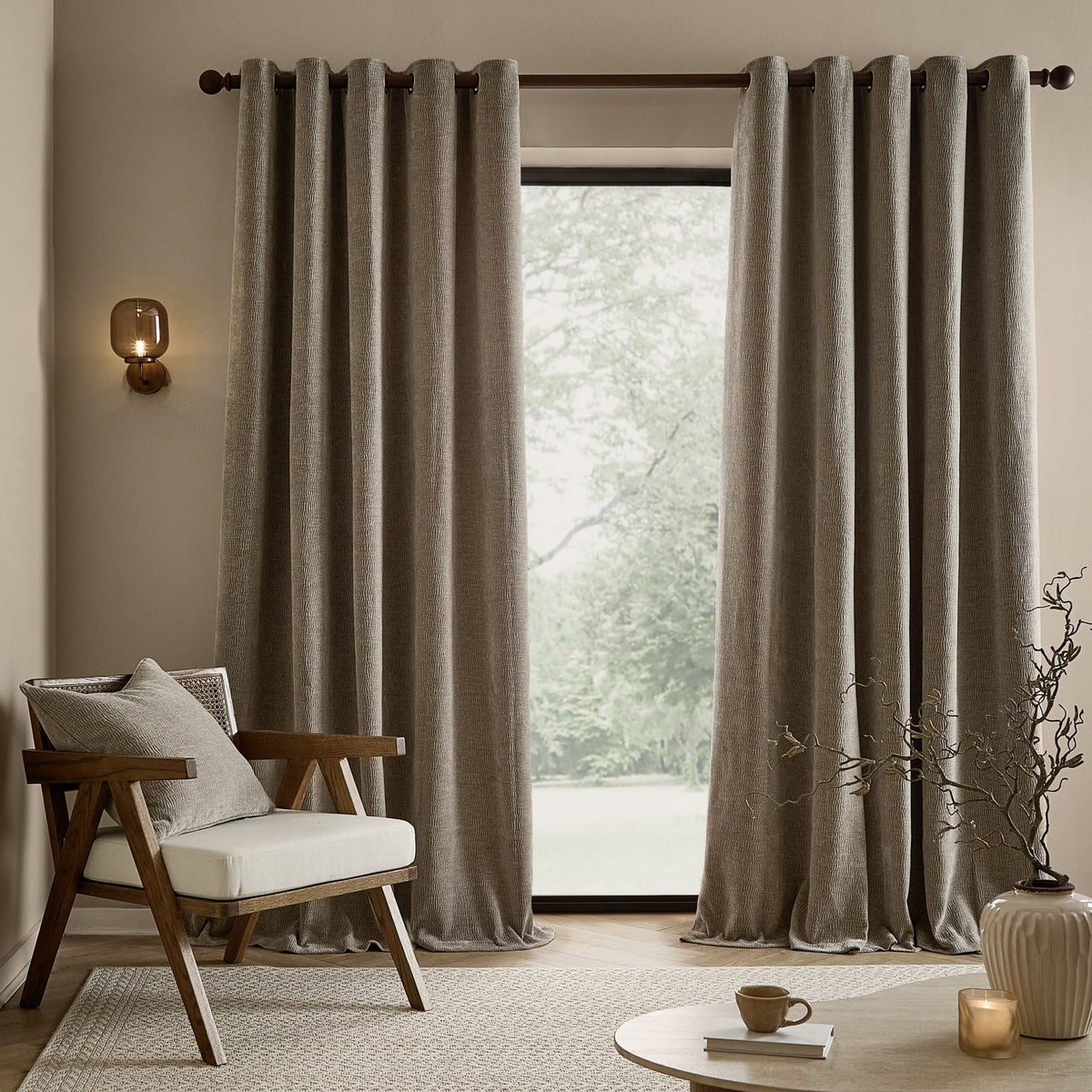 Heath Washed Velvet Jacquard Fully Lined Eyelet Curtains in greige, hung on a dark rod beside a window in a living room with a wooden armchair.