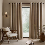Heath washed velvet linen eyelet curtains in beige hang fully lined by a window in a living room, styled with a wooden armchair, side table, and vase.