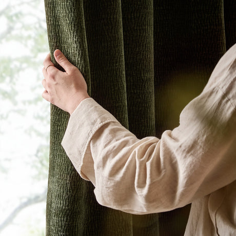 Hand drawing back an olive green Heath eyelet curtain, showing the washed velvet jacquard texture and fully lined fabric by a window.