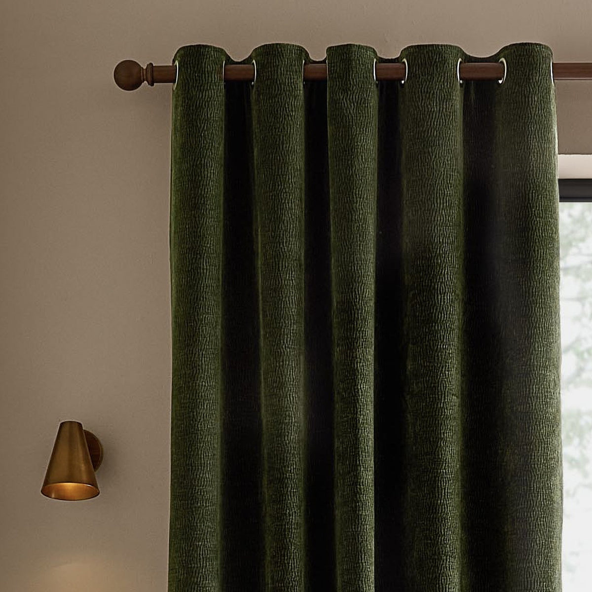 Heath eyelet curtains in olive green washed velvet with jacquard texture, shown hanging from a wooden pole with brass wall light and window nearby.