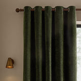 Heath eyelet curtains in olive green washed velvet with jacquard texture, shown hanging from a wooden pole with brass wall light and window nearby.