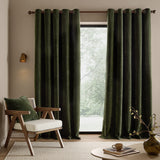 Heath washed velvet jacquard fully lined eyelet curtains in olive green hanging by a glass door, styled with a wooden armchair and matching cushion in a living room.