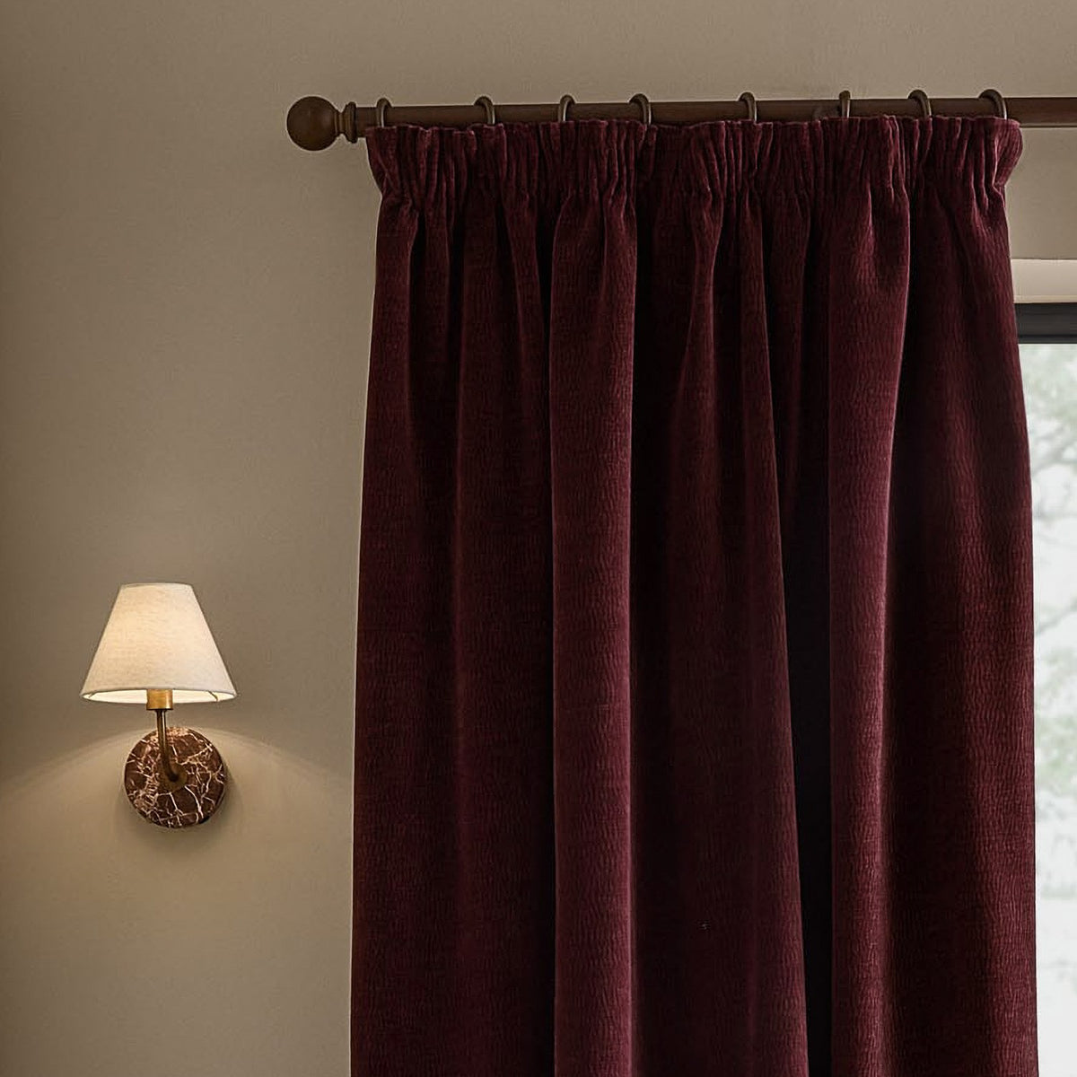 Heath Washed Velvet Pencil Pleat Curtains in beetroot hang from a wooden pole with rings, shown beside a wall-mounted lamp in a bedroom setting.
