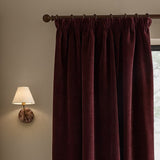 Heath Washed Velvet Pencil Pleat Curtains in beetroot hang from a wooden pole with rings, shown beside a wall-mounted lamp in a bedroom setting.