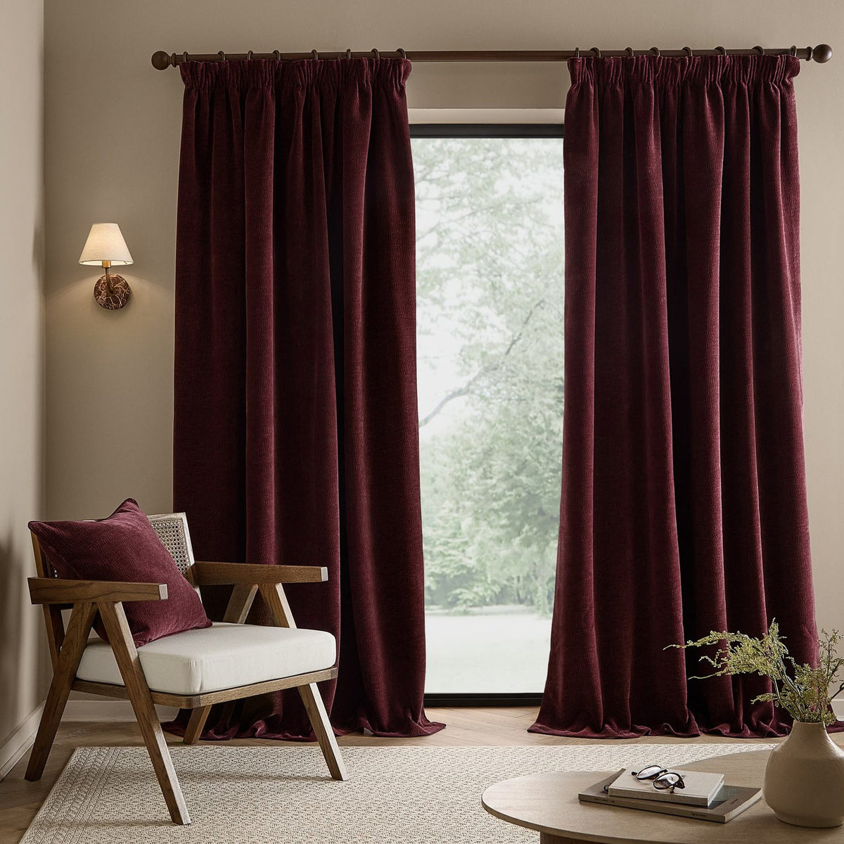 Heath Washed Velvet Pencil Pleat Curtains in deep beetroot red hang beside a window in a living room, styled with a wooden armchair and cushion.