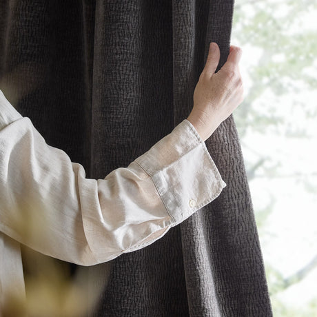 Heath Washed Velvet Pencil Pleat Curtains in charcoal, close-up of a hand in a linen sleeve holding the textured fabric beside a window.