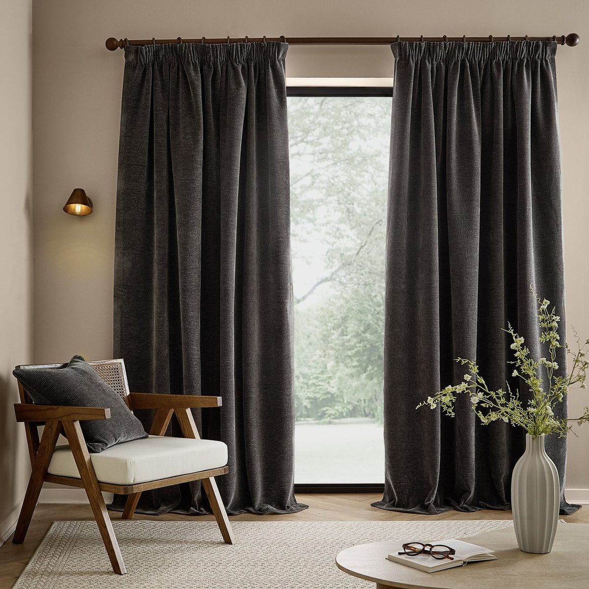 Charcoal Heath washed velvet pencil pleat curtains hanging full-length beside a glass door in a living room with a wooden armchair and vase.