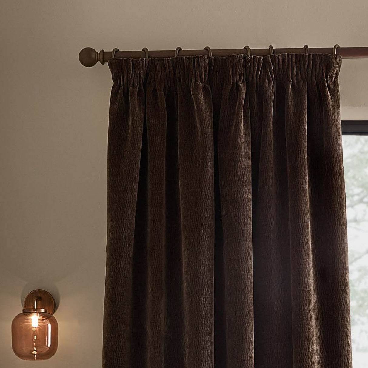 Heath washed velvet pencil pleat curtains in espresso brown, shown hanging on a bronze-toned pole with pencil pleat tape heading, in a room with a wall-mounted glass lamp.