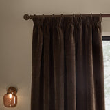 Heath washed velvet pencil pleat curtains in espresso brown, shown hanging on a bronze-toned pole with pencil pleat tape heading, in a room with a wall-mounted glass lamp.