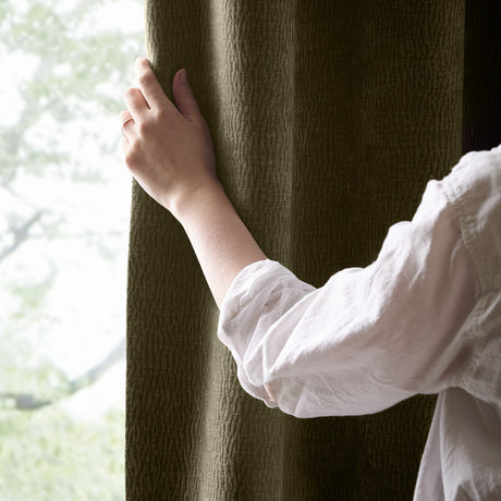 Hand in a white shirt reaching for the Heath pencil pleat curtains in Eucalyptus, showing the washed velvet texture and olive green colour by a window.