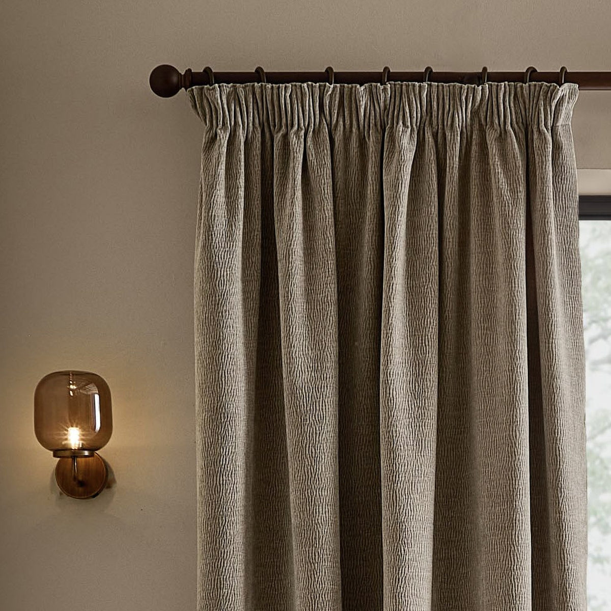 Heath washed velvet pencil pleat curtains in greige, hung on a dark rod with pencil pleat heading, shown beside a window in a softly lit room with a wall-mounted glass sconce.