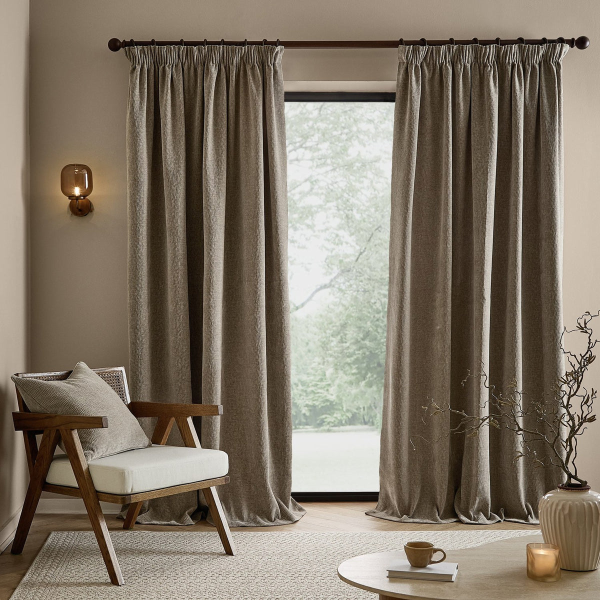 Heath washed velvet pencil pleat curtains in greige, shown floor-length either side of a window in a living room with an armchair, coffee table and wall light.