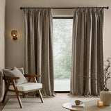Heath washed velvet pencil pleat curtains in greige, shown floor-length either side of a window in a living room with an armchair, coffee table and wall light.