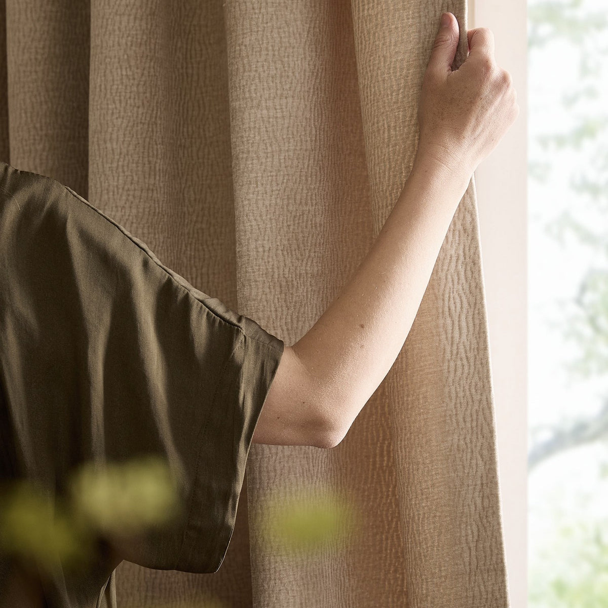 Heath Washed Velvet Eyelet Linen Pencil Pleat Curtain in beige, shown by a window with a hand gathering the textured jacquard fabric to the side.