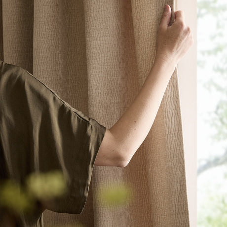 Heath Washed Velvet Eyelet Linen Pencil Pleat Curtain in beige, shown by a window with a hand gathering the textured jacquard fabric to the side.