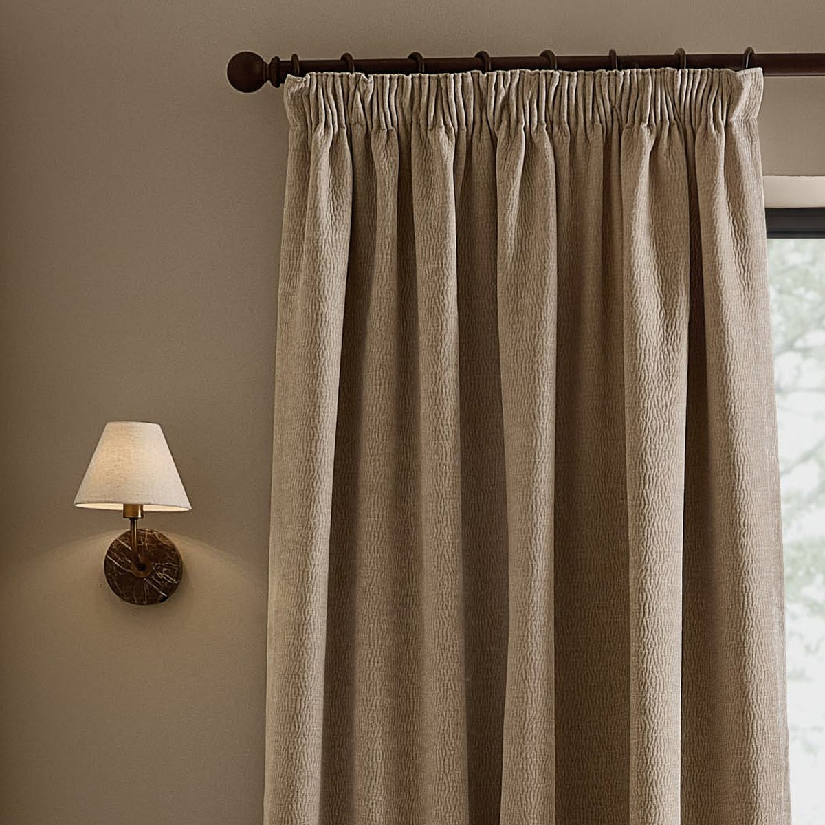 Heath Washed Velvet Pencil Pleat Curtains in linen beige, textured jacquard fabric with gathered heading on a dark rod, shown beside a wall-mounted lamp.