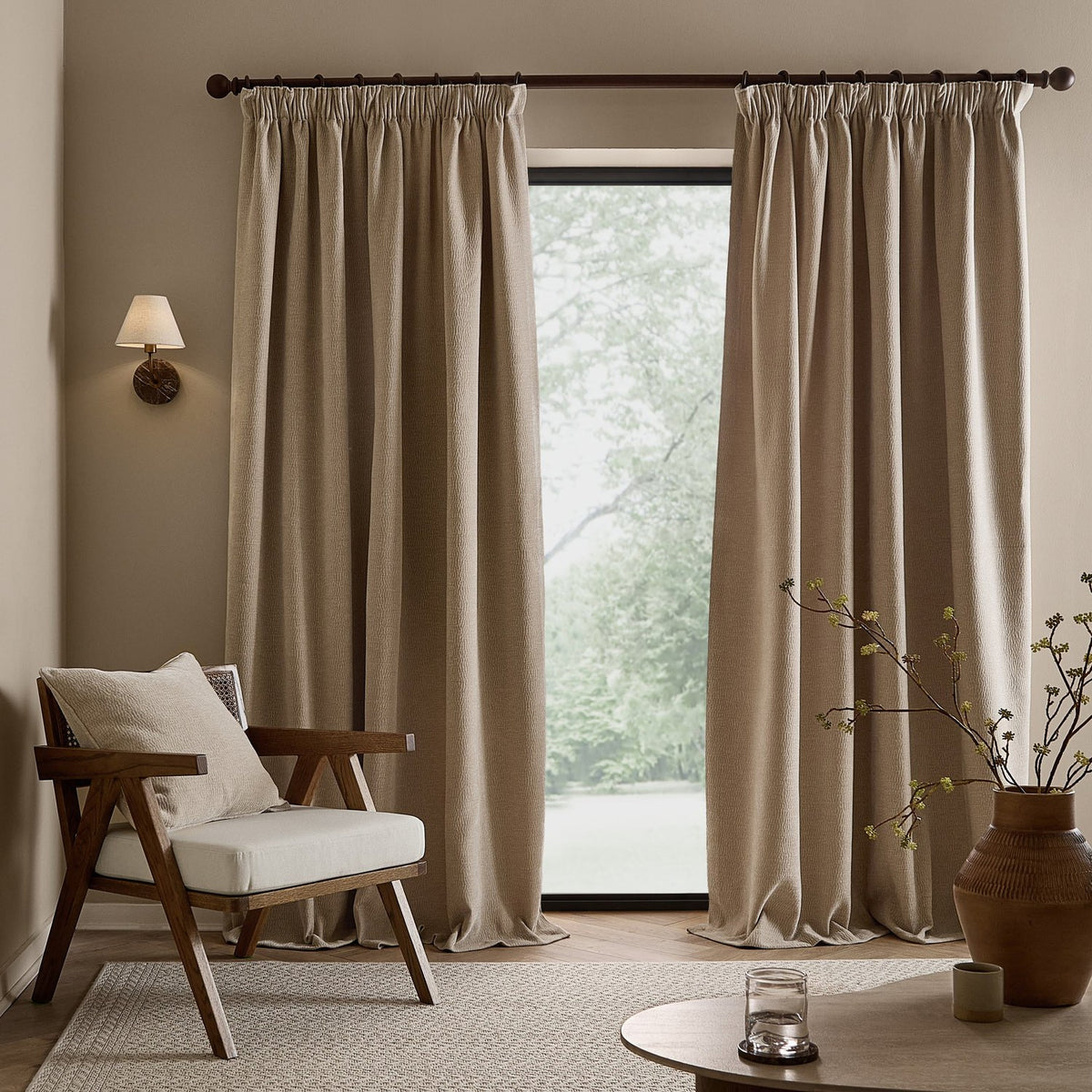 Heath Washed Velvet Pencil Pleat Curtains in linen beige, shown floor-length beside a window in a living room with an armchair, rug, and side table.