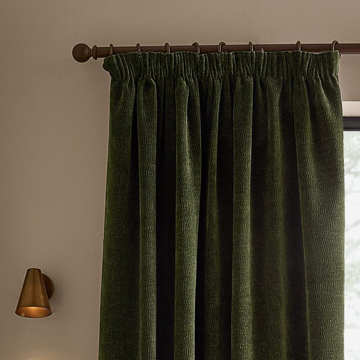 Heath Washed Velvet Pencil Pleat Curtains in olive green, shown with pencil pleat heading on a dark wood pole with rings, hanging in a living room beside a brass wall sconce.