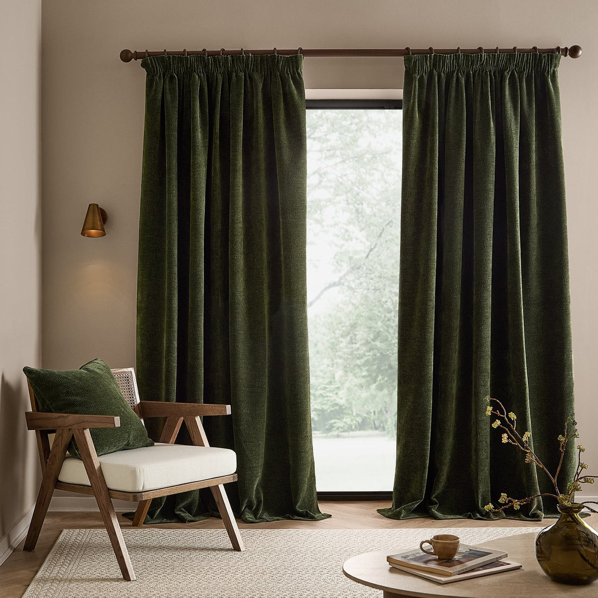 Heath washed velvet pencil pleat curtains in olive green hanging full-length by a window, shown in a living room with a wooden armchair and round coffee table.
